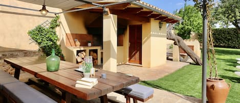 Een terras buiten met een houten tafel en banken, een pergola en een grote terracotta pot.