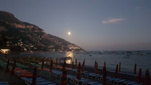 Ein Strand mit Liegestühlen und Sonnenschirmen, ein ruhiges Meer mit Booten und ein Berg im Hintergrund.