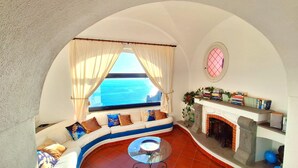 Une pièce avec un canapé incurvé, une table basse en verre, une cheminée et une vue sur la mer par une fenêtre ornée de rideaux.