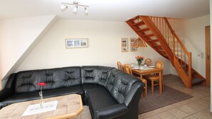 A living room with a black leather sofa, a wooden dining table with chairs, a wooden staircase, and a ceiling light fixture. - Seeschlößchen Thatched Cottage, "Sturmmöwe" Vacation Rental, just 200 meters from the beach (Büsumer Deichhausen)