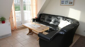 A living room with a black leather sofa, a wooden coffee table, a potted plant, and a sliding glass door leading to a balcony. - Seeschlößchen Thatched Cottage, "Sturmmöwe" Vacation Rental, just 200 meters from the beach (Büsumer Deichhausen)