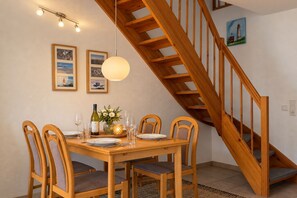 A dining area with a wooden table set for four, surrounded by chairs with cushioned seats. A wooden staircase with a railing is visible in the background.