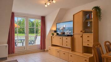 A room with a television on a wooden cabinet, a dining table with chairs, and a view through a glass door to an outdoor area.