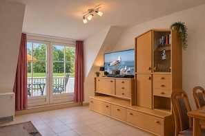 A room with a television on a wooden cabinet, a dining table with chairs, and a view through a glass door to an outdoor area.