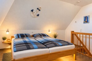 A bedroom with a sloped ceiling, a bed with a checkered blanket, two bedside lamps, a small plant, and a painting of a lighthouse.