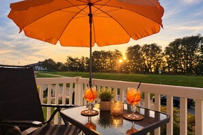 A table with drinks and a potted plant on a balcony with a white railing, overlooking a grassy area with trees and a sunset in the background.