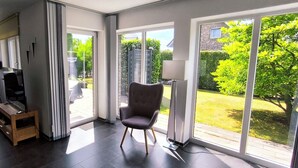 A modern living room with a large sliding glass door leading to a garden. There are two armchairs, a wooden side table, and a floor lamp. - Fantastic holiday home on the left Lower Rhine between the Rhine and Wisseler See (Kalkar)