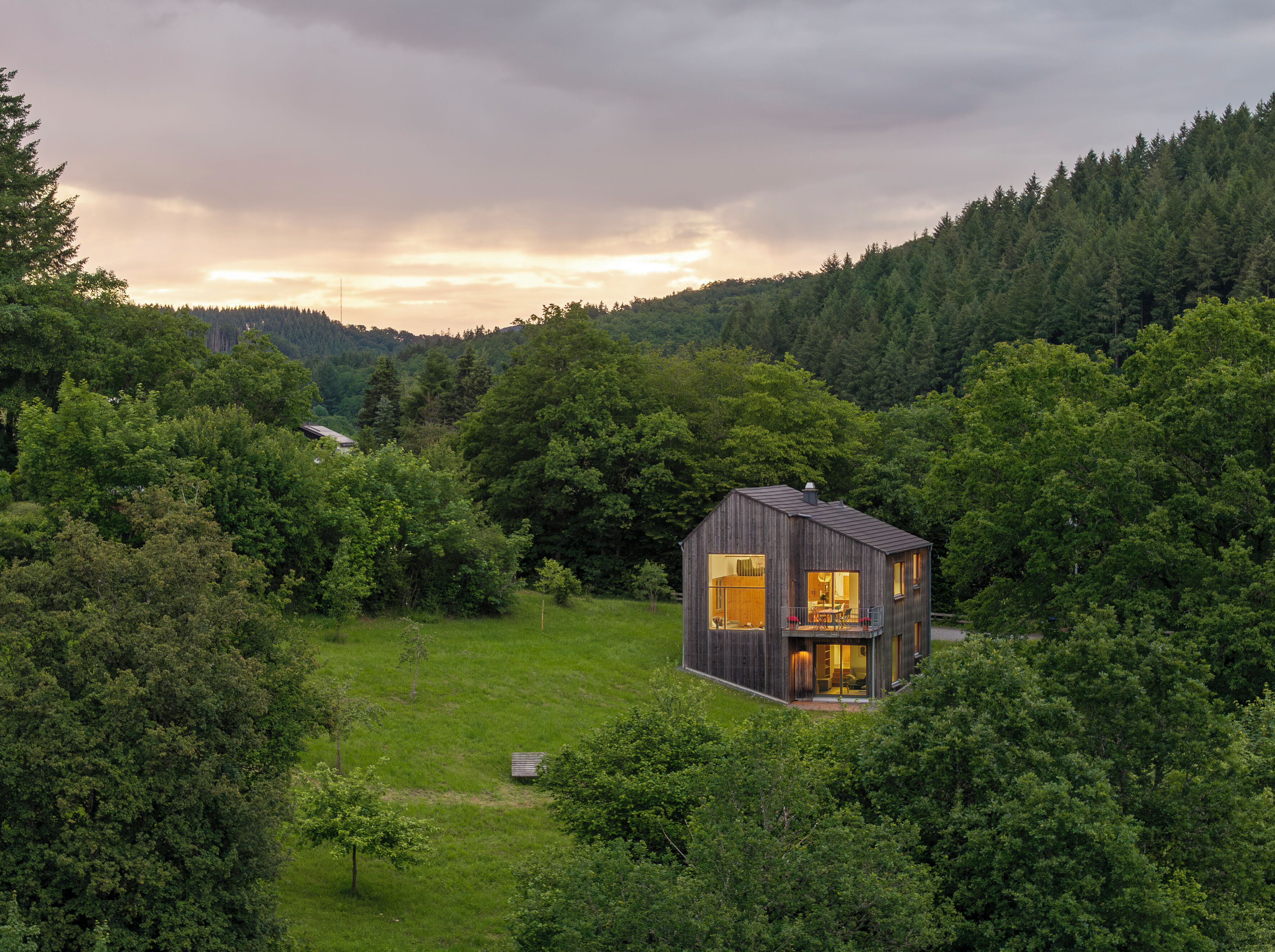 A modern wooden cabin with large windows, surrounded by dense forest, set on a grassy hill.