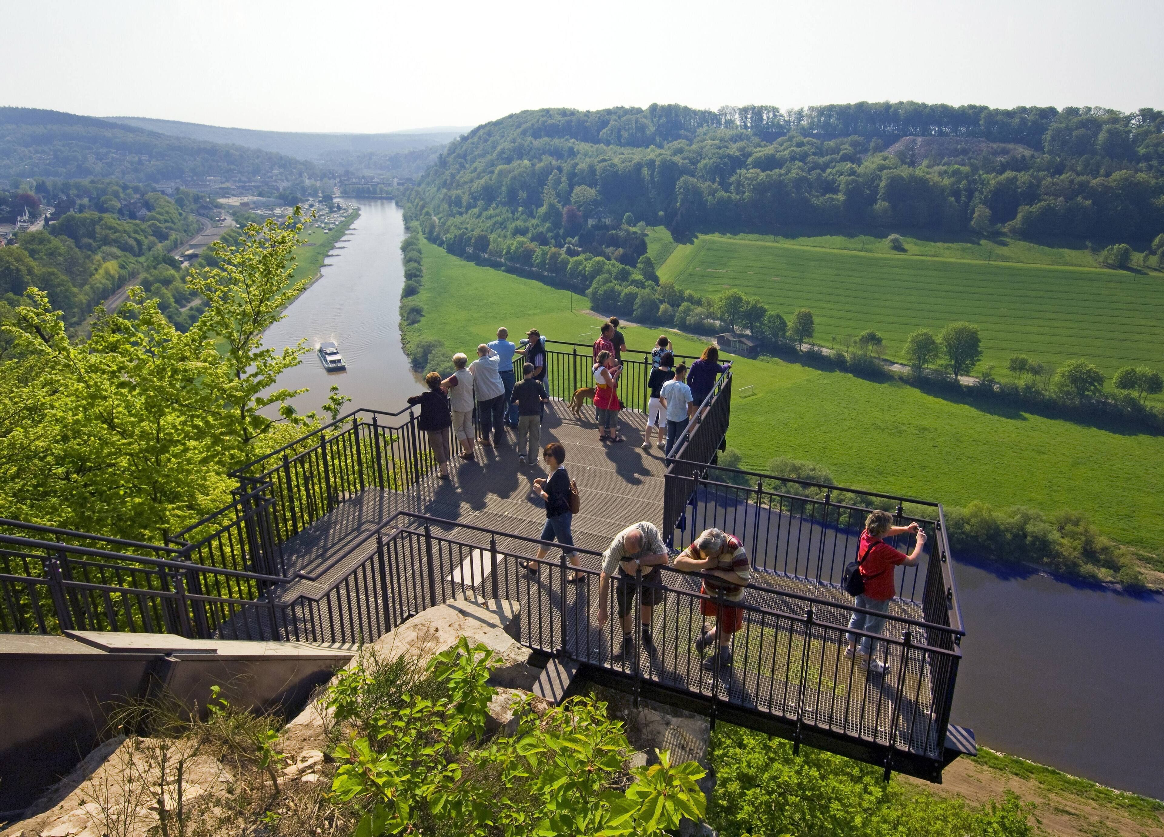 Un belvédère offrant une vue panoramique sur une rivière et de vastes champs verdoyants, avec une rambarde en métal et des escaliers menant à la rive.