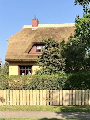 Ein traditionelles Haus mit einem Strohdach und Schornstein, umgeben von üppiger Vegetation und einem Holzzaun.
