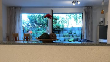 Un coin repas avec vue sur un jardin, grâce à une grande fenêtre, une table en bois agrémentée d’un vase de fleurs, et un espace cuisine équipé d’une machine à café.