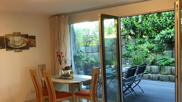En moderne stue med en sort sofa, et spisebord med stole og en skydedør, der fører ud til en udendørs terrasse med bord og stole.