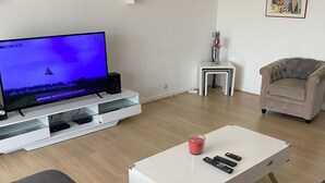 Ein modernes Wohnzimmer mit einem Flachbildfernseher auf einem weißen Schrank, einem weißen Couchtisch mit Fernbedienungen, einem grauen Sofa, einem beigen Sessel und einem kleinen weißen Beistelltisch mit einer Kerze.