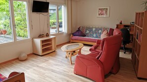 A living room with a sofa, a red chair, a small round table, a TV mounted on the wall, and a bookshelf.