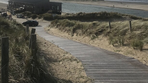 A beachside wooden boardwalk leading to a building with a flat roof, situated on sandy dunes with sparse vegetation. - House for 5 persons, old farmhouse with luxe bathroom, 3 sleeping rooms (Groet)