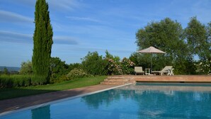 Una piscina con un cielo azzurro limpido, un albero alto e una zona relax con sedie e un ombrellone.