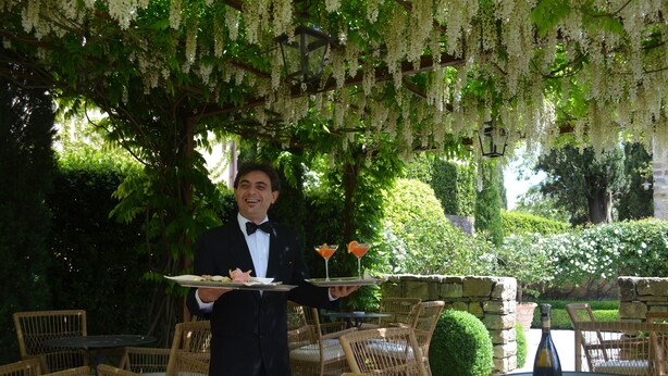 Un cameriere che porta un vassoio con un piatto di cibo e due bevande, in un giardino con mobili in vimini e fiori bianchi pendenti.