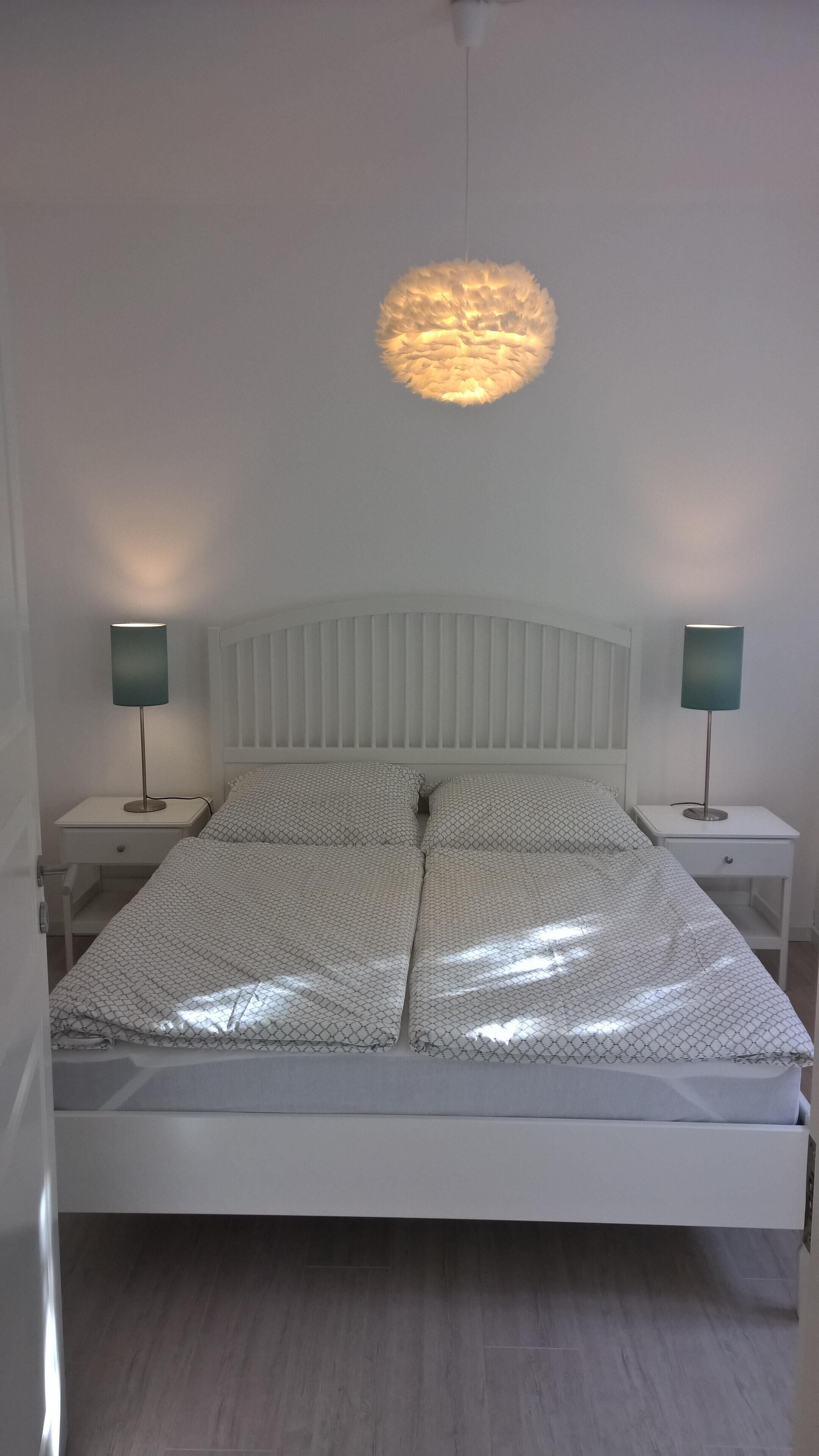 Een slaapkamer met een wit bed, twee nachtkastjes met lampen, een wit hoofdeinde en een hanglamp.