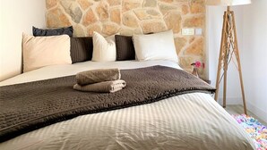 A bedroom with a stone wall, a bed with a striped duvet, multiple pillows, a bedside lamp, and a floor lamp. - Centrally located in Pollensa with wonderful roof terrace views (Pollença)