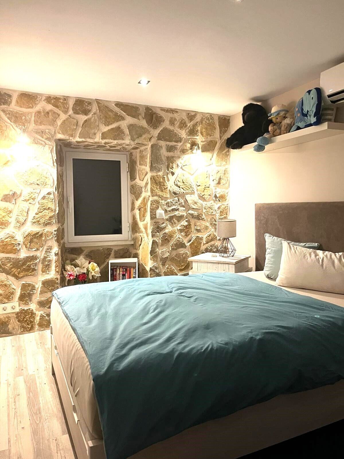 Et soveværelse med en stenvæg, en seng med et blåt lagen, et natbord med en lampe og en hylde med bamser.