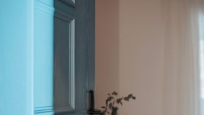 A room with a wooden floor, a blue door, a small wooden table with a plant, and a white bed.