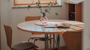 A dining area with a round table, chairs, a hanging light fixture, a window with blinds, and a small plant on the windowsill.