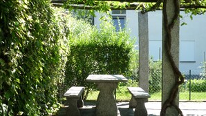 Mesa e bancos de pedra em área ajardinada, com cerca e vegetação.