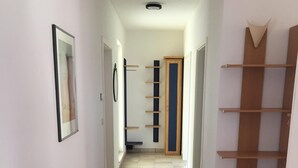 Un couloir étroit au sol carrelé, avec un tableau encadré sur le mur de gauche, une porte avec une étagère verticale en bois sur le mur de droite, et une porte au fond.