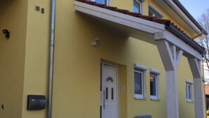 Un bâtiment jaune avec une porte blanche, un petit porche avec un banc et le numéro « 10 B » inscrit sur le mur.