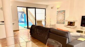 Un salon moderne avec un canapé gris, des fauteuils orange, une table basse en verre, une cheminée et une vue sur une piscine et un escalier.