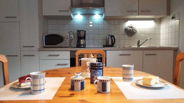 A kitchen with a wooden table set for breakfast, including a plate of cake, a coffee pot, a kettle, and several mugs.