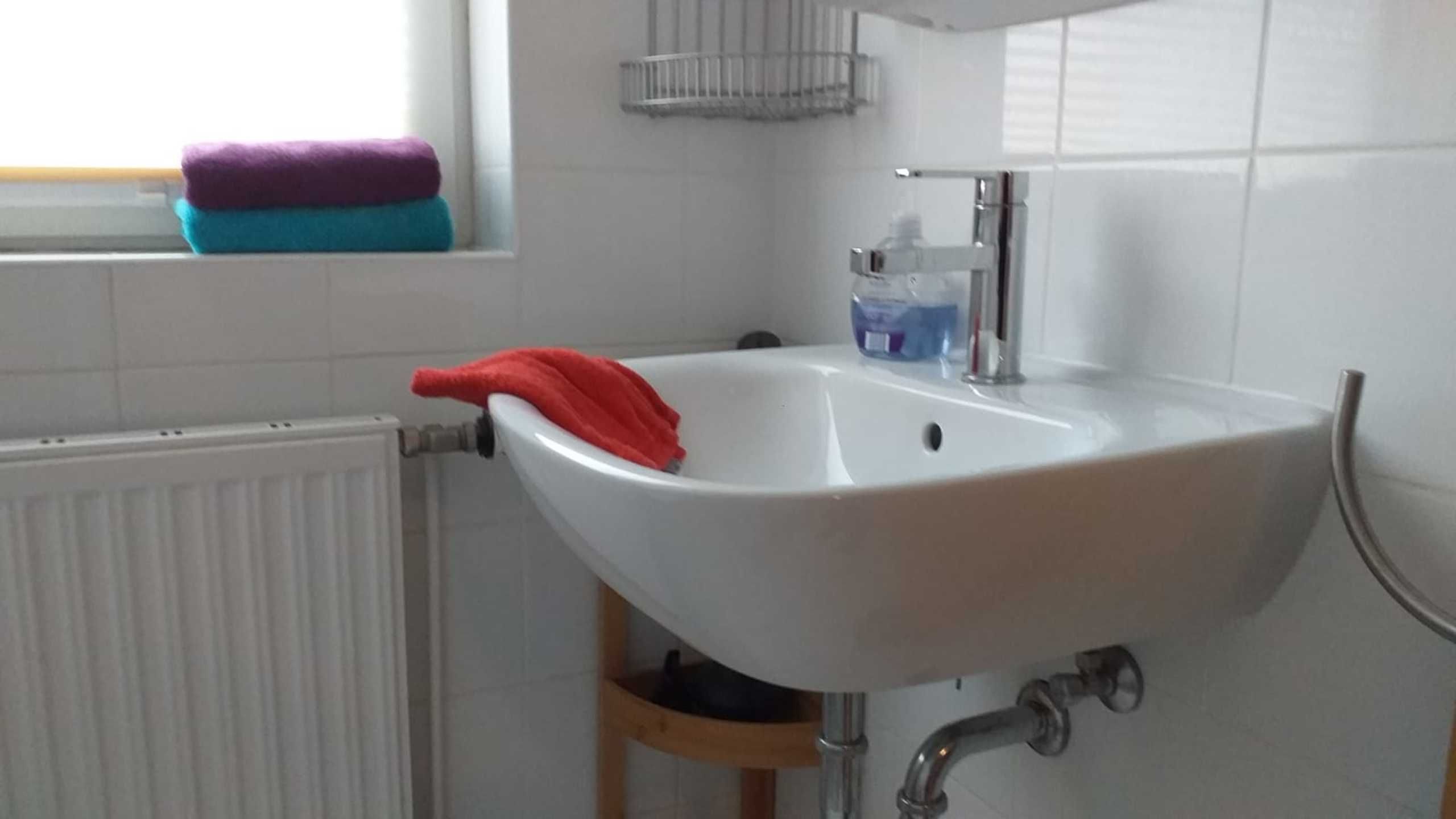Ein weißes Waschbecken mit einem roten Handtuch, ein weißer Heizkörper und ein Fenster mit einem blau-violetten Handtuch.