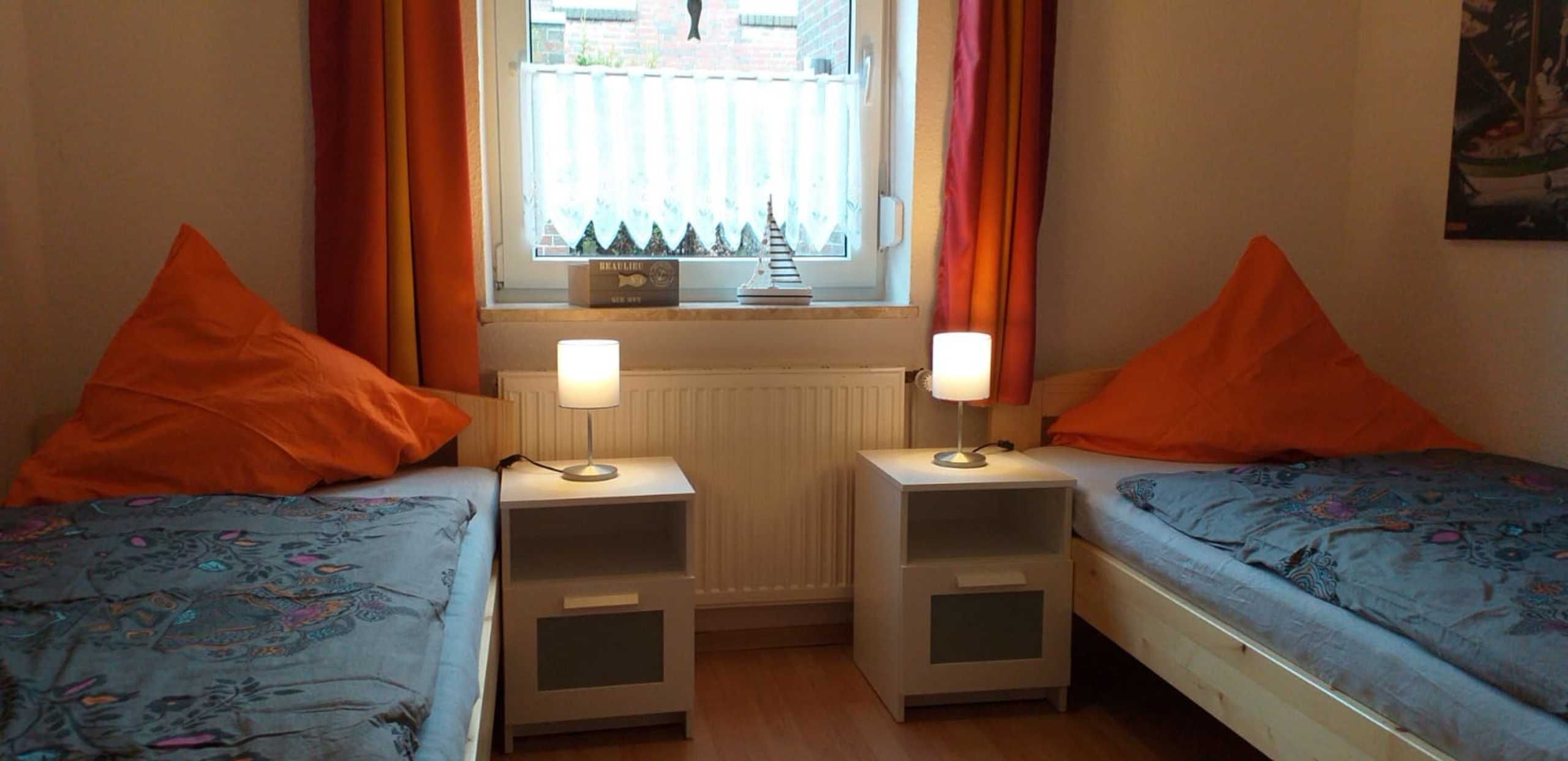 Una habitación con dos camas, cada una con un cojín naranja y una mesita de noche con lámpara. Se observa una ventana con cortinas y un radiador encima.