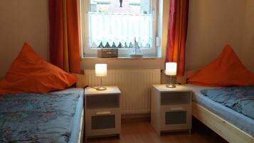 Una habitación con dos camas, cada una con un cojín naranja y una mesita de noche con lámpara. Se observa una ventana con cortinas y un radiador encima.