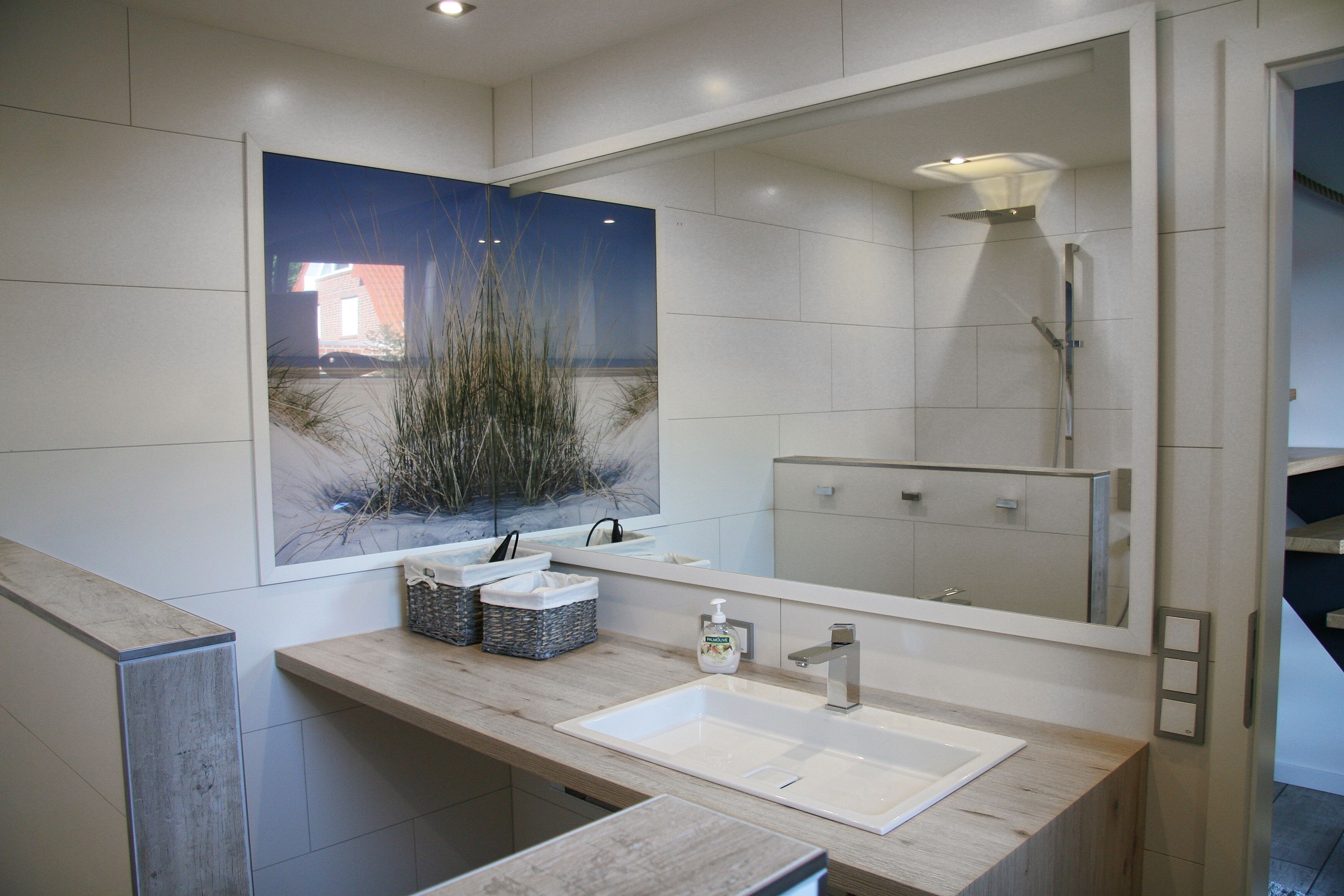 Ein modernes Badezimmer mit einem großen Spiegel, einem Waschbecken und einer Dusche. Auf der Ablage liegen Körbe und ein Fenster bietet Blick auf ein Gebäude und Grünflächen.