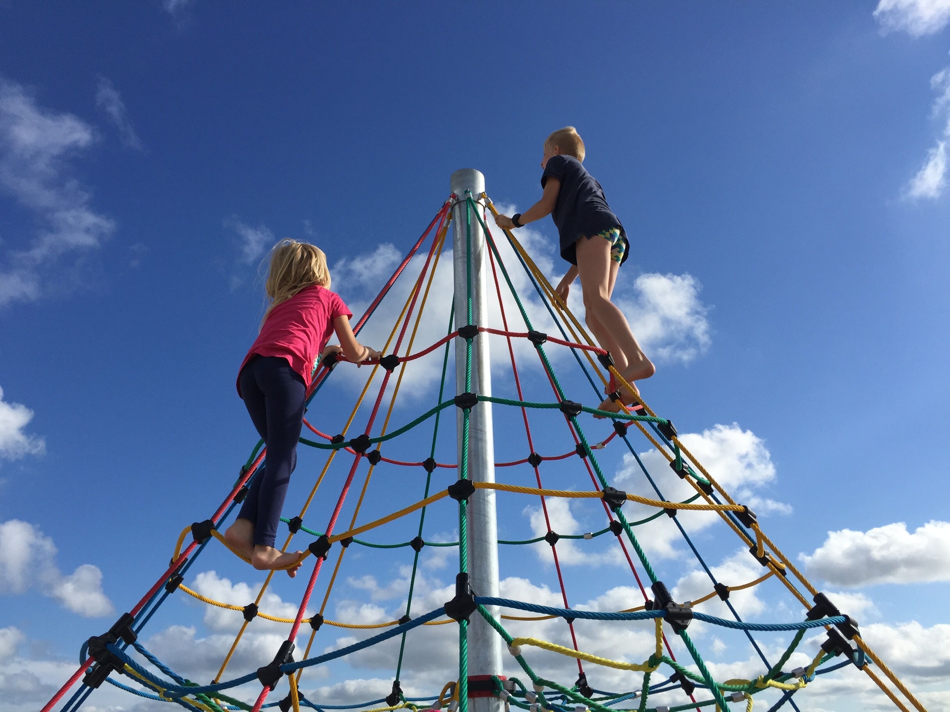 Two children climbing a rope net structure against a clear blue sky.
