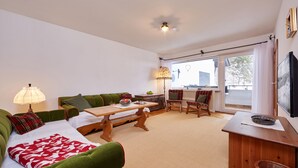 A cozy living room with a green sofa, a wooden table, and a chair. There is a lamp, a window with curtains, and a TV on the wall. - Apartment on the Zugspitze (Grainau)