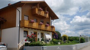 A two-story wooden house with a balcony, a car parked in front, and a garden with various plants and flowers. - Vacation on the farm in beautiful Koesslarn in Lower Bavaria (Kößlarn)