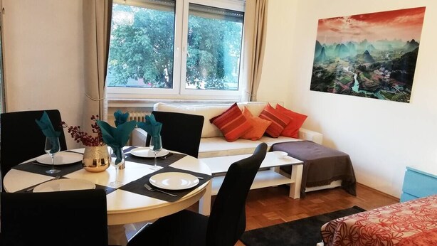Un comedor con una mesa redonda preparada para cuatro, un sofá con cojines rojos y un cuadro en la pared.