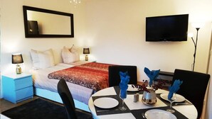 Una habitación de hotel con una cama, un televisor, una mesa de comedor con platos y servilletas, y una mesita auxiliar con una lámpara.