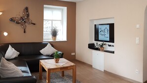 A living room with a black leather sofa, a wooden coffee table, a decorative butterfly wall art, a television mounted on the wall, and a window with a plant on the windowsill. - Holiday home for sole use, garden sauna, outdoor whirlpool, WLAN, play barn (Plütscheid)