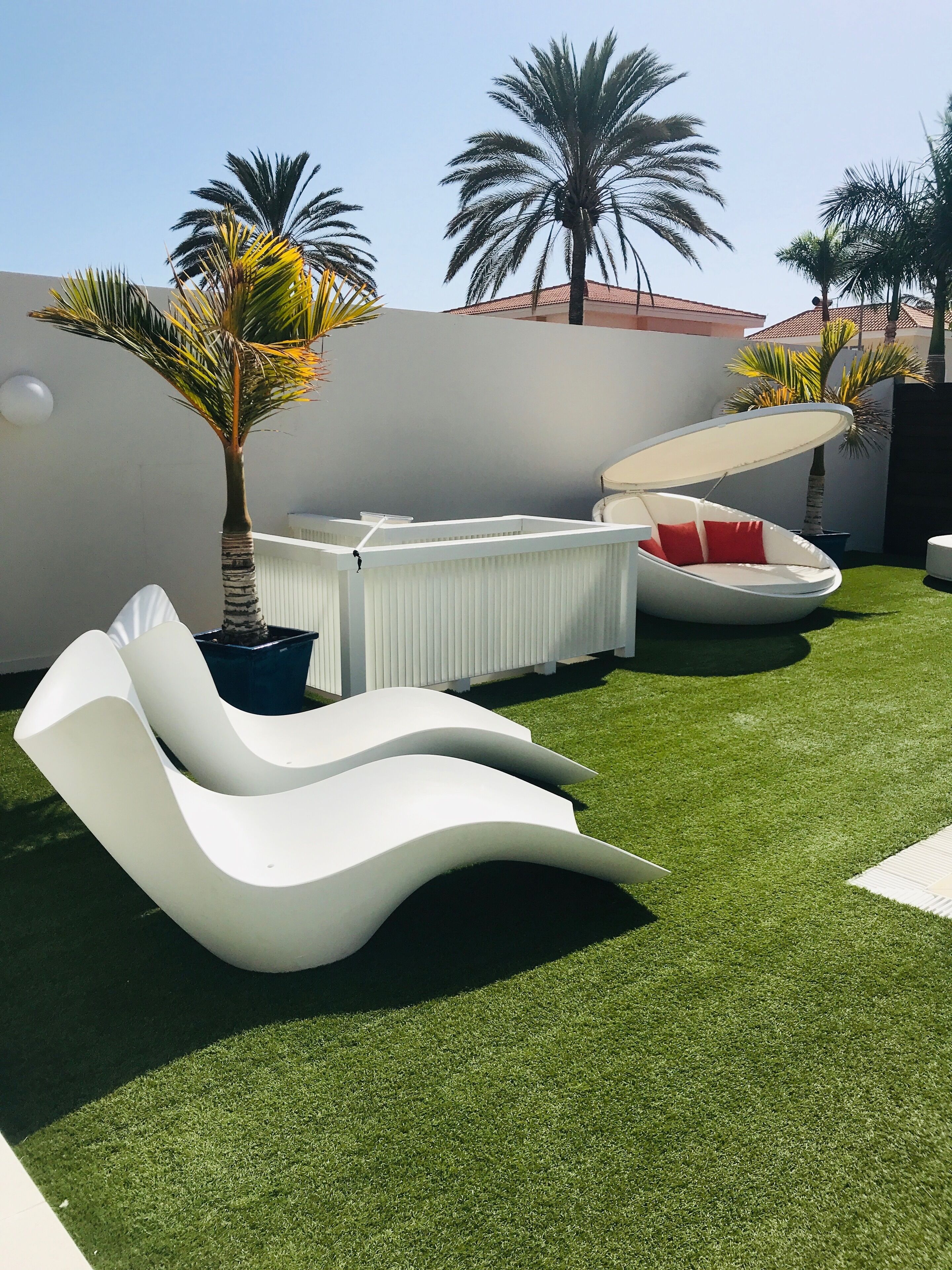 A modern outdoor seating area with white lounge chairs and a covered seating pod, surrounded by artificial grass and palm trees.