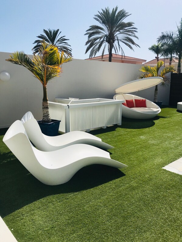 Un espace extérieur moderne, aménagé avec des fauteuils blancs et un espace de détente couvert, le tout entouré de gazon synthétique et de palmiers.
