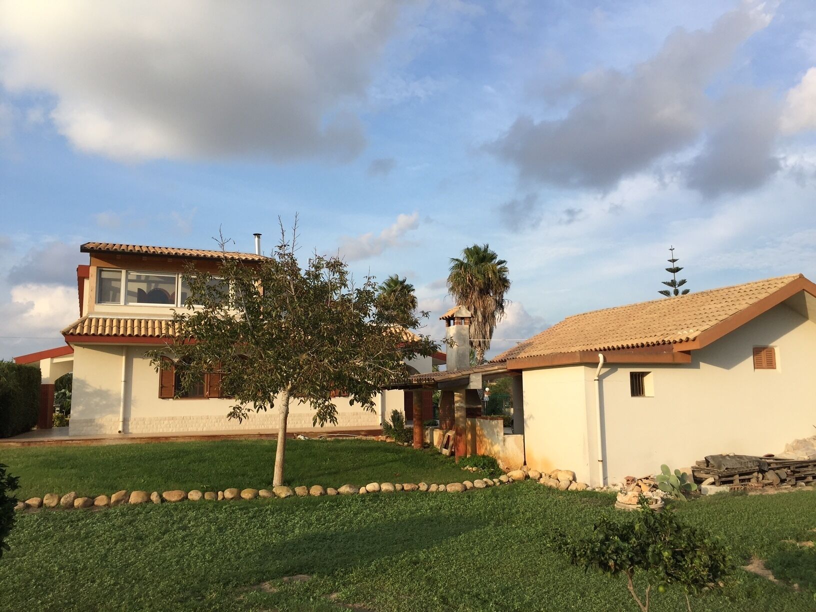 Una casa de dos pisos con techo de tejas, un pequeño jardín con un árbol y una palmera al fondo.