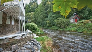 Ein rustikales Steinhaus mit einem Balkon mit Blick auf einen Fluss, umgeben von üppiger Vegetation und einer kleinen Struktur mit rotem Dach.