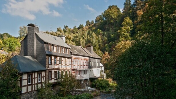 Ein traditionelles Fachwerkhaus mit Stein Fundament, gelegen an einem Bach in einem bewaldeten Gebiet.