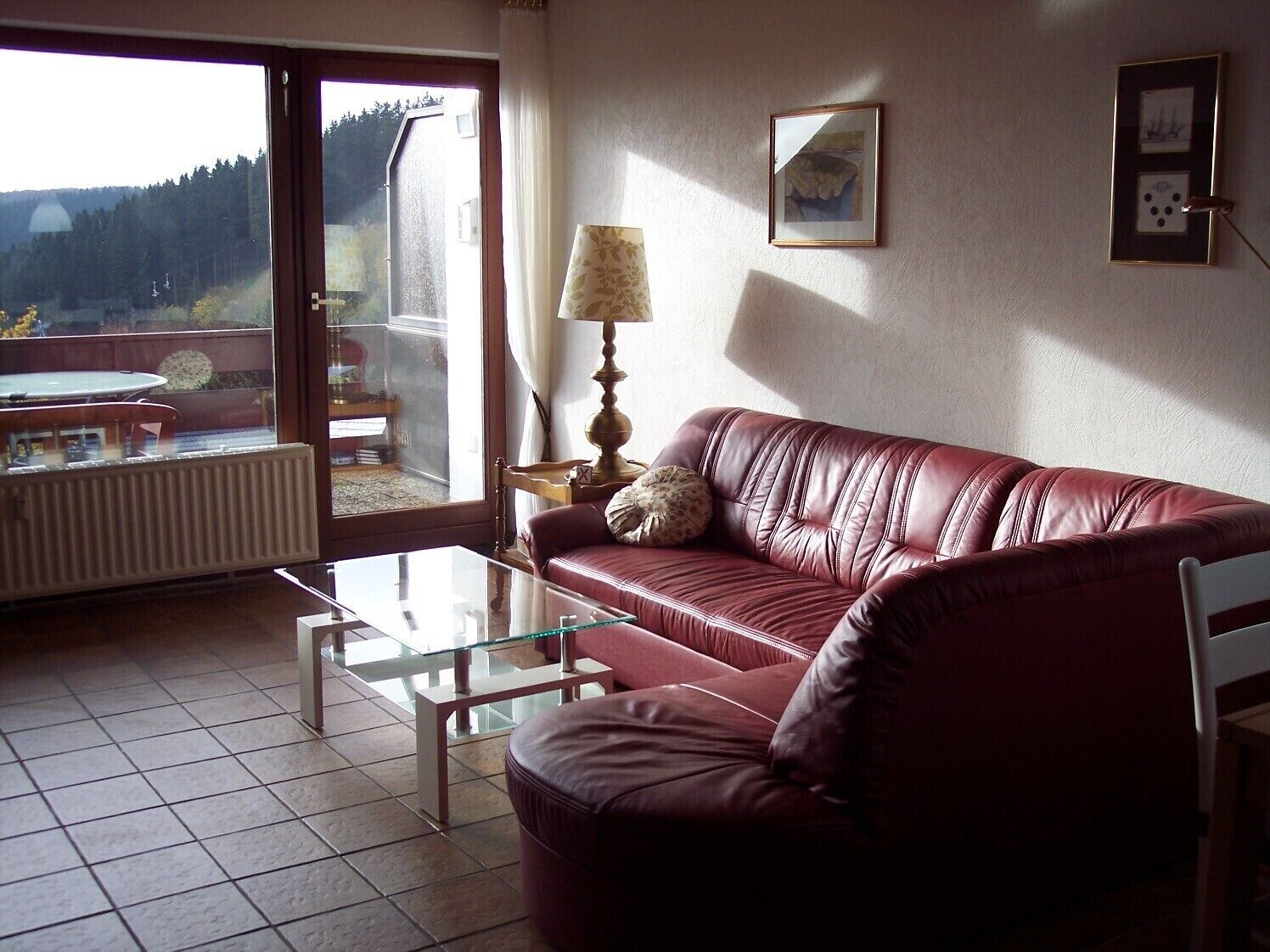 A living room with a leather sofa, a glass coffee table, a lamp, and a view of a forest through a large window.