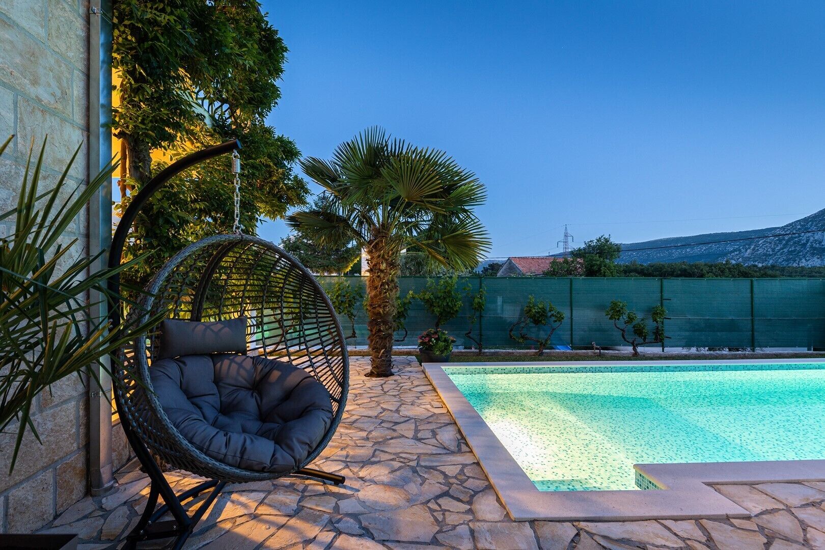 Ett poolområde med en terrass belagd med stengolv, en flätstol med kuddar och en palm.