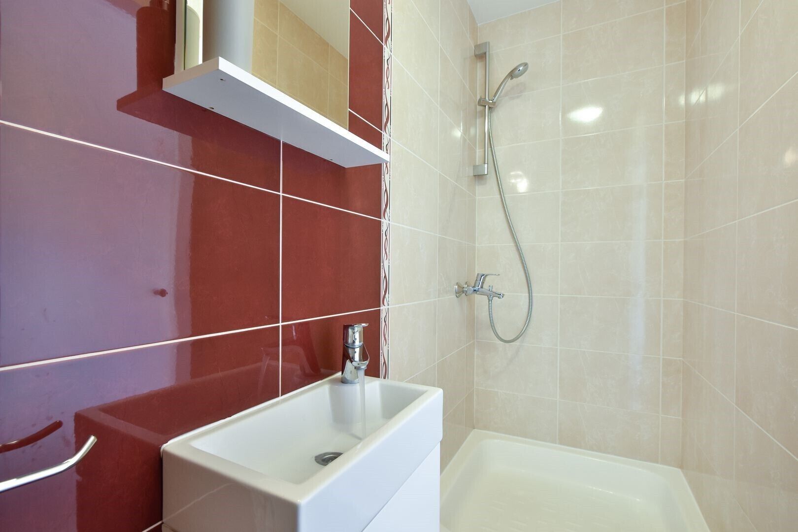 A modern bathroom with a white sink, a shower with a handheld showerhead, and red and beige tiled walls.