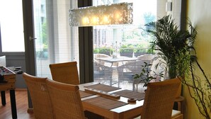Un coin repas avec une table en bois et des chaises en osier, de grandes fenêtres équipées de stores et un luminaire suspendu.